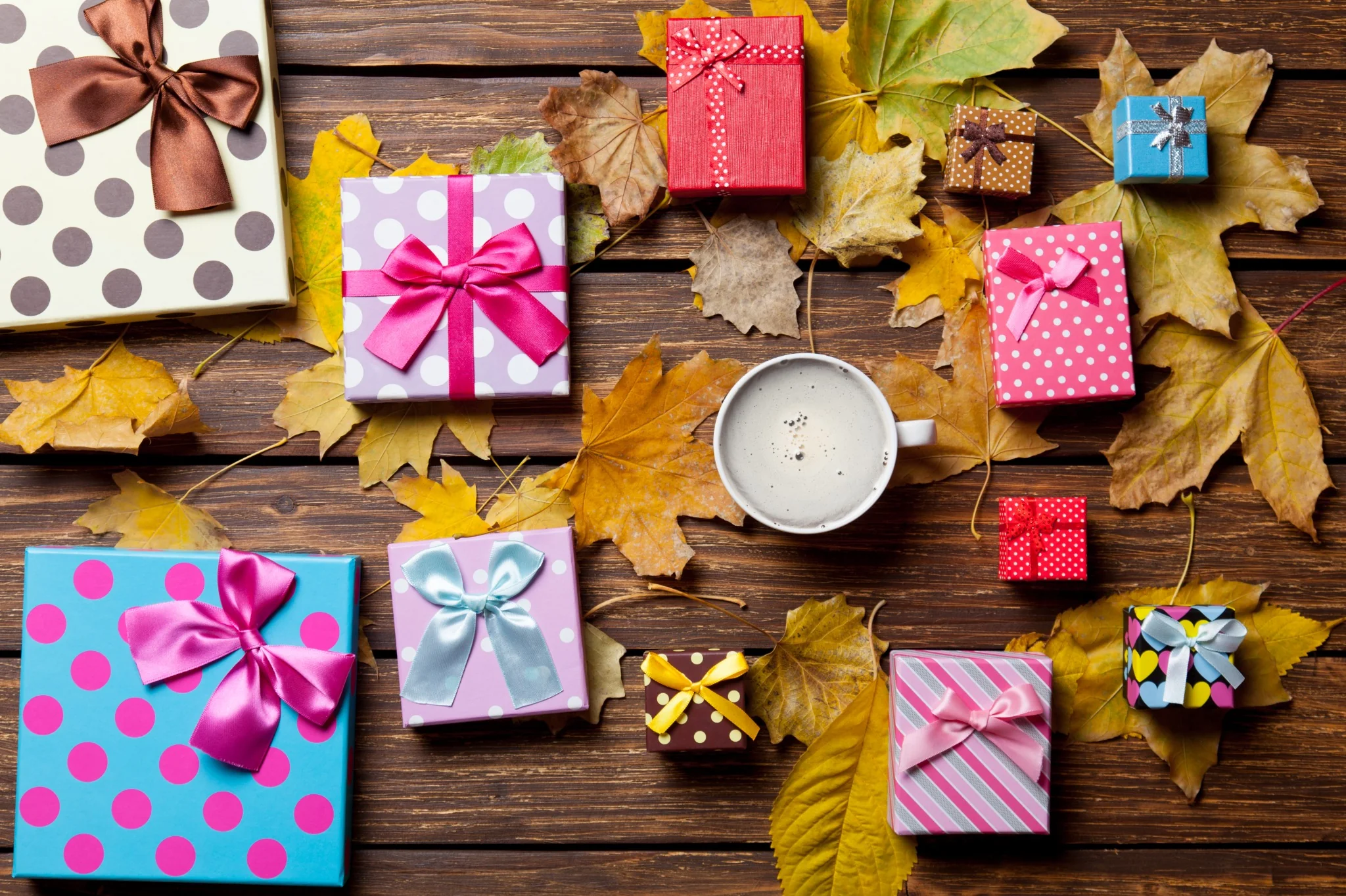 Colorfully wrapped holiday gifts with ribbons arranged among autumn leaves on a wooden table.