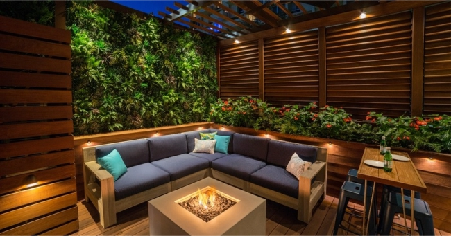 A stylish outdoor seating area at night features a built-in L-shaped sofa with accent pillows, a modern fire pit, and Coastal Source lighting.