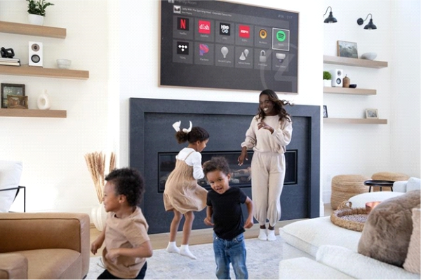 A family dancing in a living room with whole-home audio and a wall-mounted screen showing a smart home interface above the fireplace.
