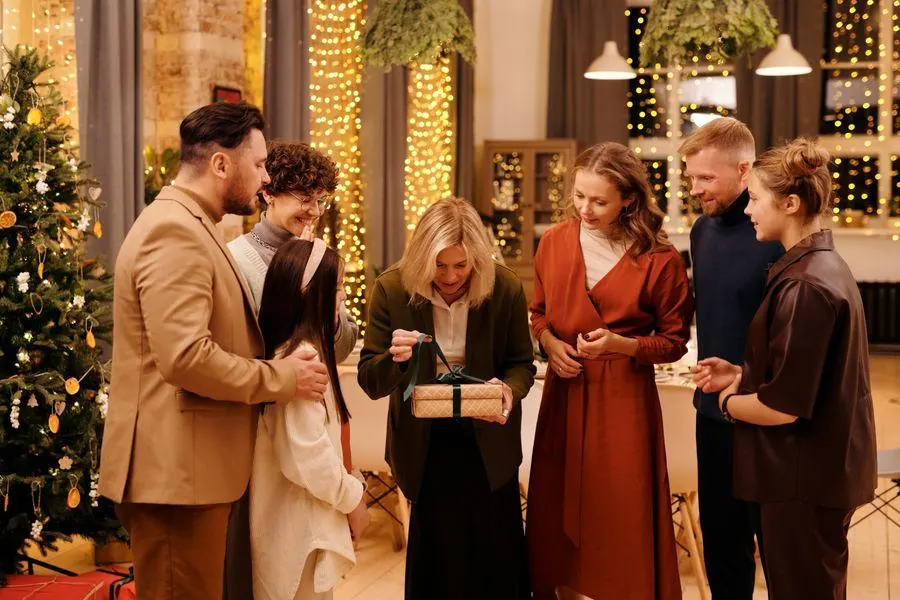 Family and friends exchanging gifts beside a decorated Christmas tree in a warmly lit home.