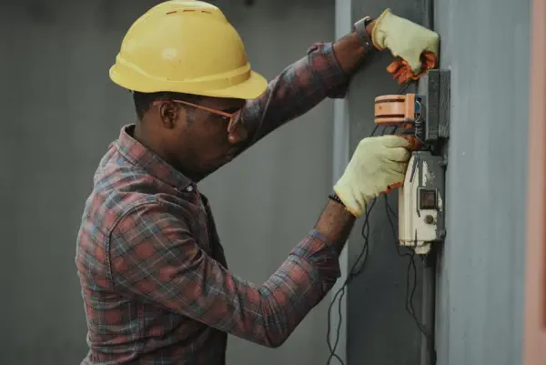An electrician wearing a yellow hard hat, gloves, and safety glasses installs wiring into an exterior electrical box on a building wall.