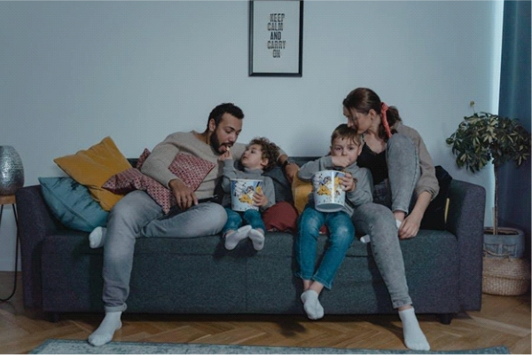 A family of four sitting together on a couch, enjoying popcorn while watching something, creating a cozy and relaxed home entertainment atmosphere.