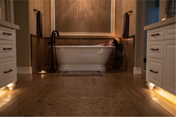 Woman relaxes in a bathtub surrounded by soft accent lighting in a luxury bathroom with fully lowered shades.