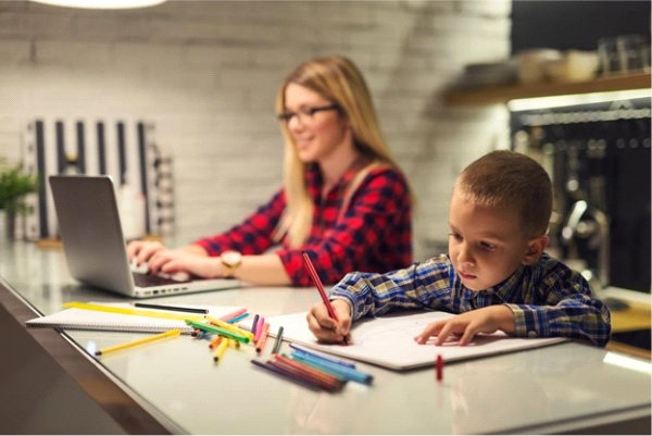 Child drawing with colored pencils at a kitchen counter while an adult works on a laptop in the background.