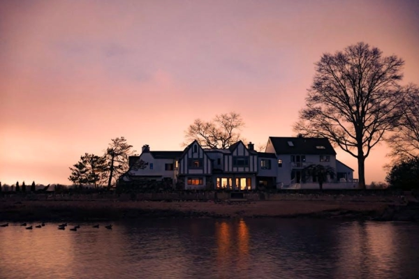 Large waterfront home silhouetted against a pink and orange sunset with interior lights reflecting on the calm water.