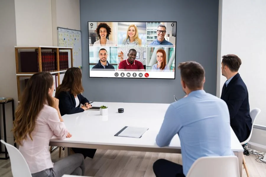 Employees in a modern conference room participating in a video call with remote colleagues displayed on a large wall-mounted screen.