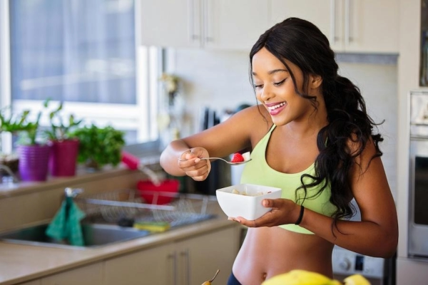 A woman in exercise clothes smiling and eating fruit in the kitchen.